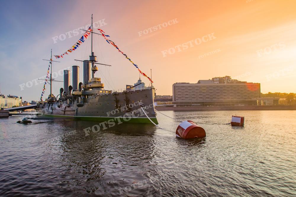 На фотографии изображен вид на реку и крейсер "Аврора" в Санкт-Петербурге, украшенный праздничными флагами. Закатное небо переливается теплыми оттенками, освещая водную гладь и добавляя сцене волшебства. Изображение может быть использовано для образовательных материалов, исторических исследований, туристической рекламы или как элемент дизайна, вдохновляющий на путешествия и изучение культурного наследия.
